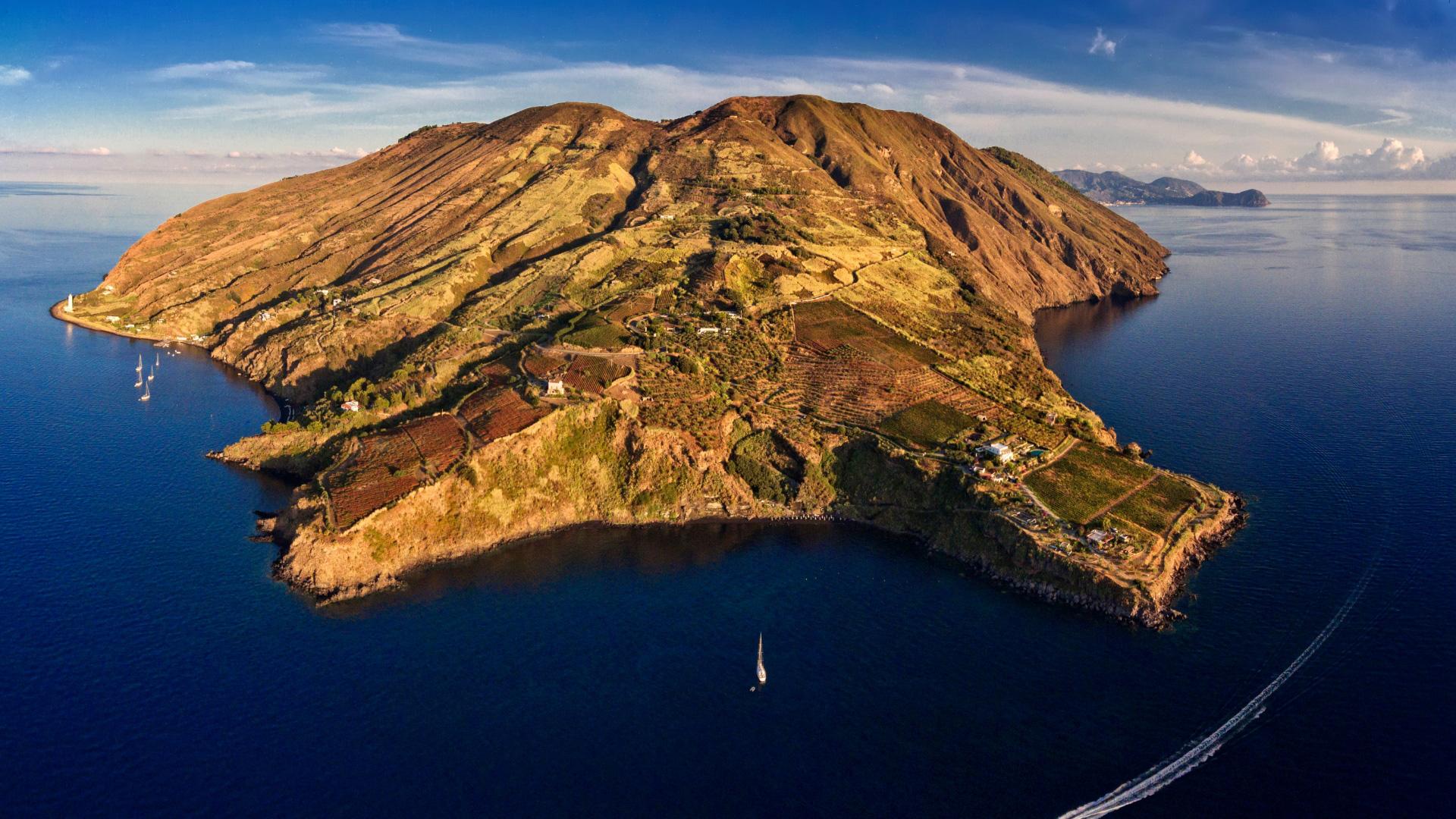 Isola vulcanica con coste rocciose e campi coltivati, circondata dal mare.