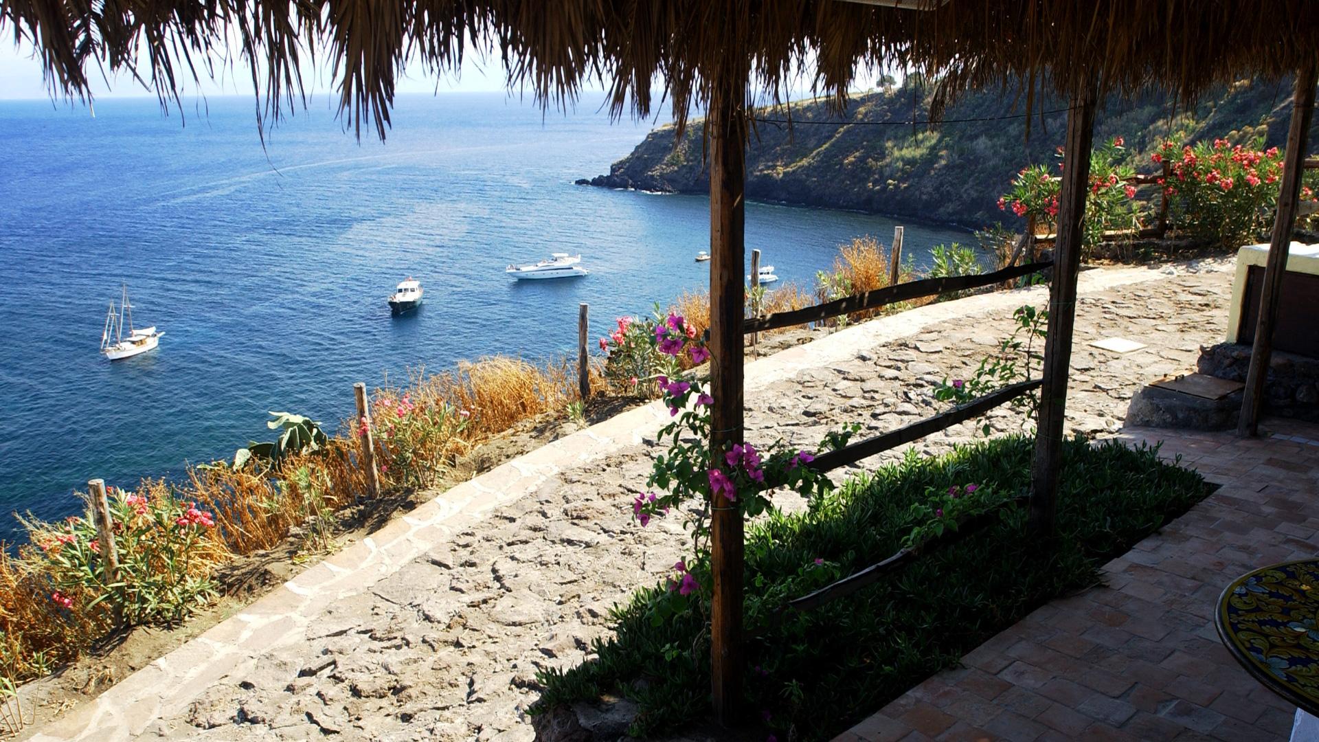 Vista panoramica sul mare con barche, fiori e sentiero in pietra.