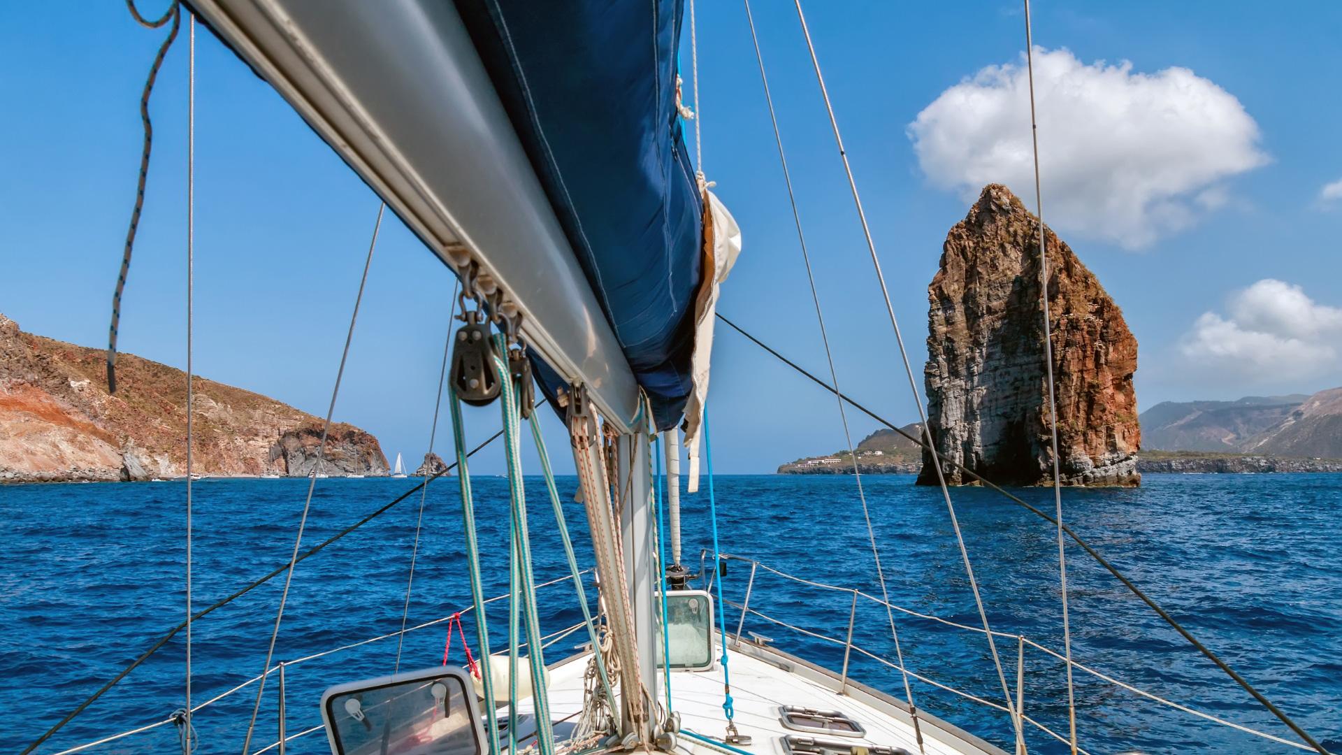 Barca a vela naviga vicino a un imponente faraglione in mare aperto.